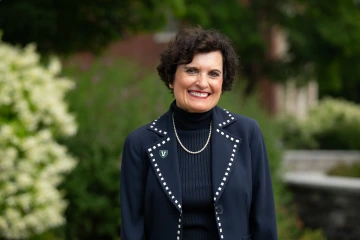 Patricia Prelock standing outside with green bushes and a brick building behind her.